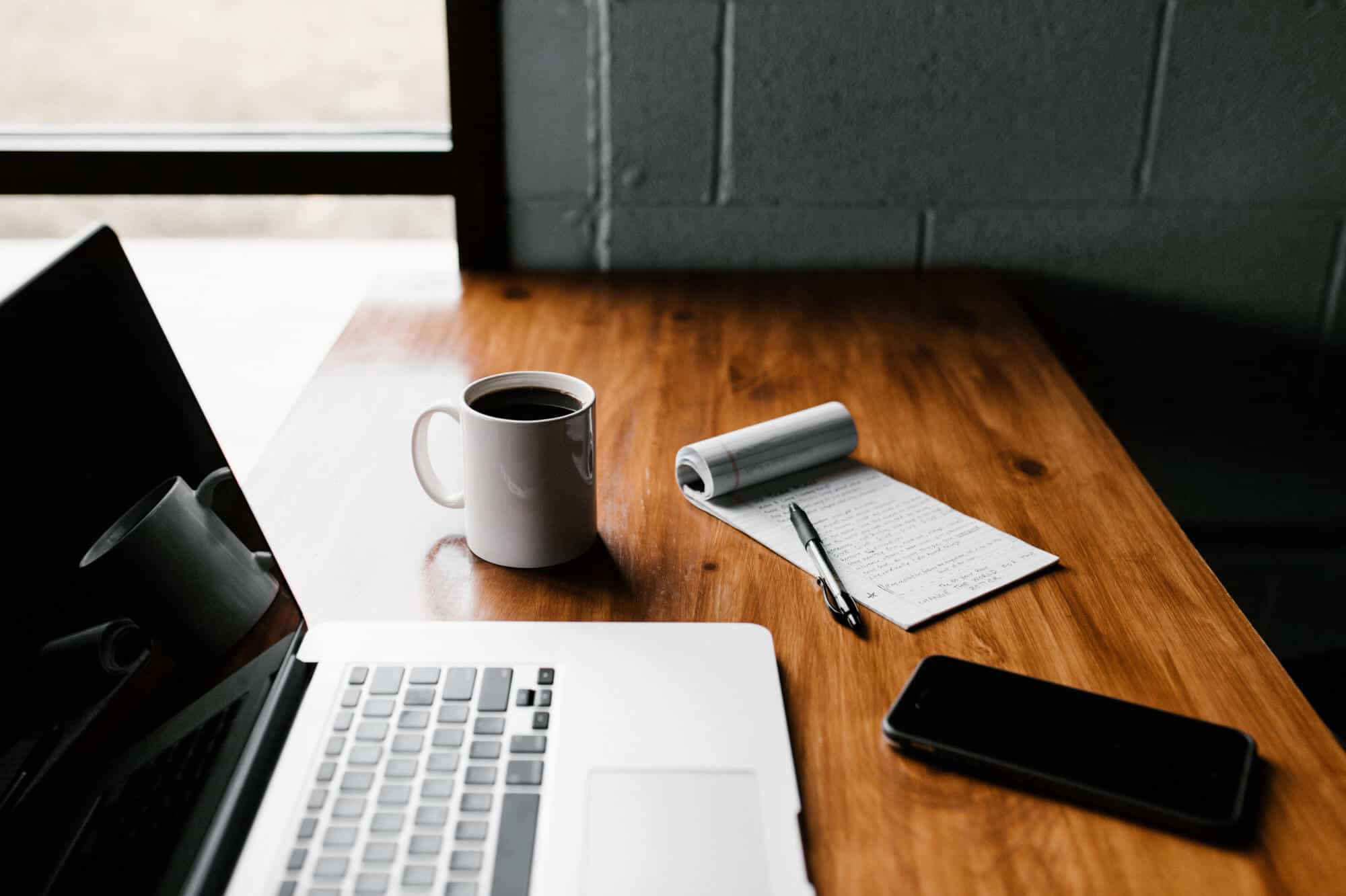 A laptop, notebook and phone laid out on a desk beside a cup of coffee