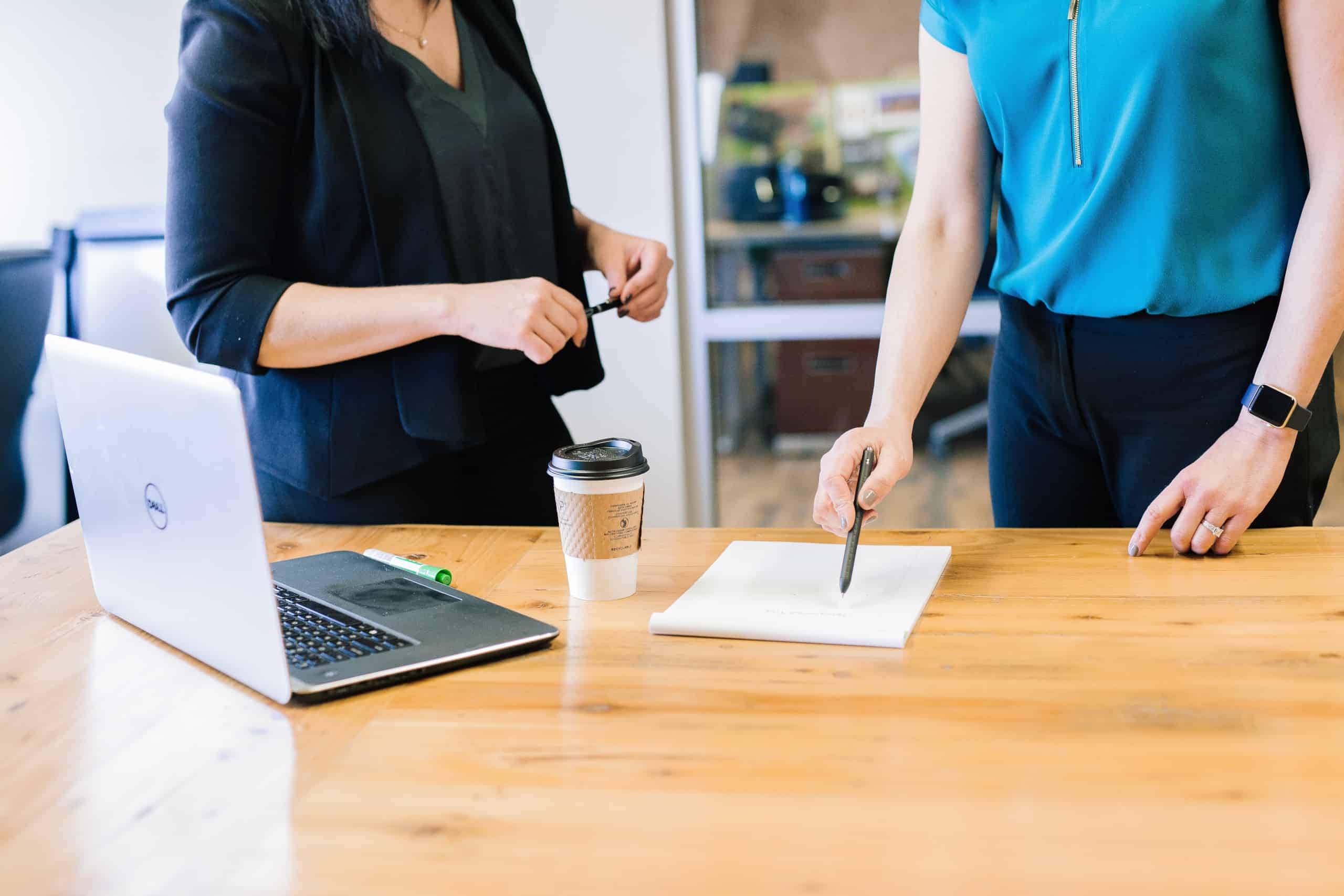 Two people at a desk pointing at a notepad and signing paperwork beside an open laptop and a coffee cup