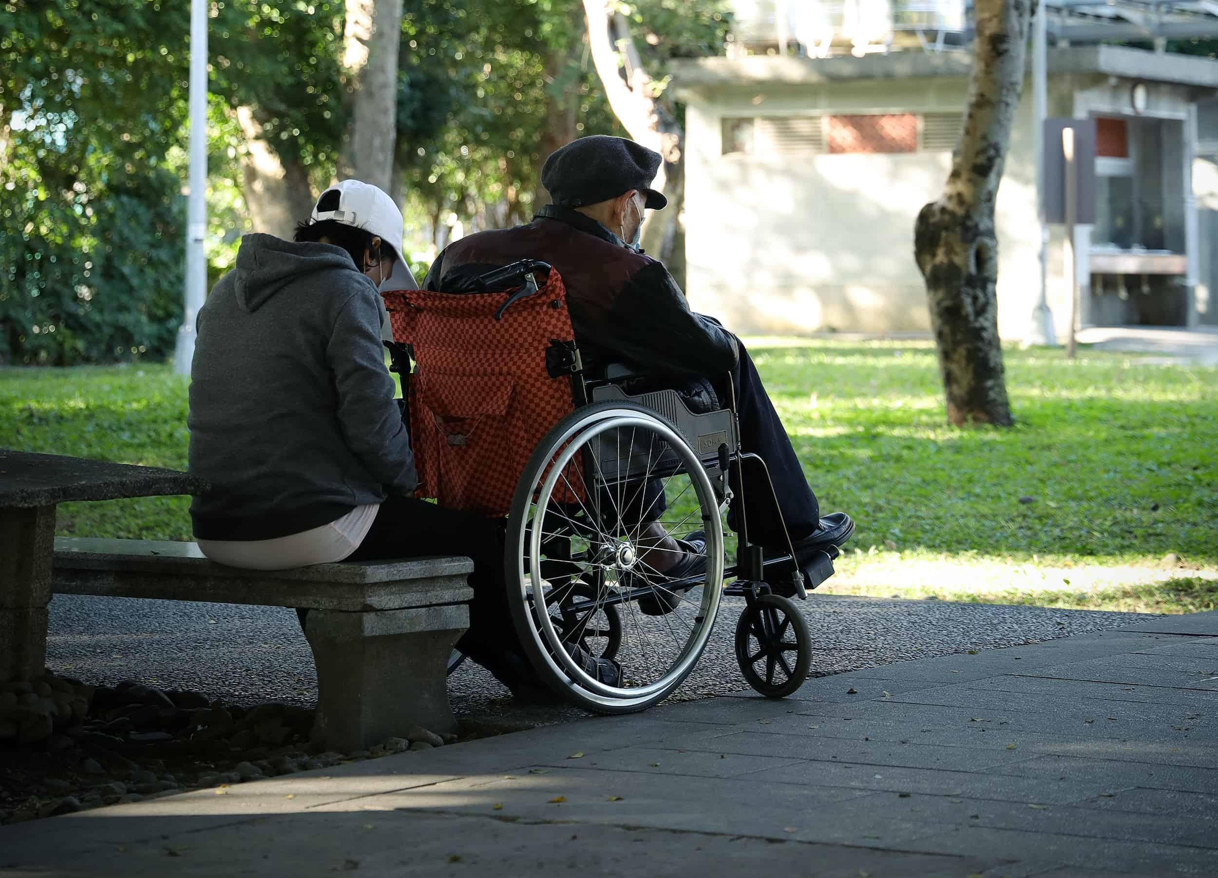 A carer sitting on a bench beside her client's wheelchair in a park