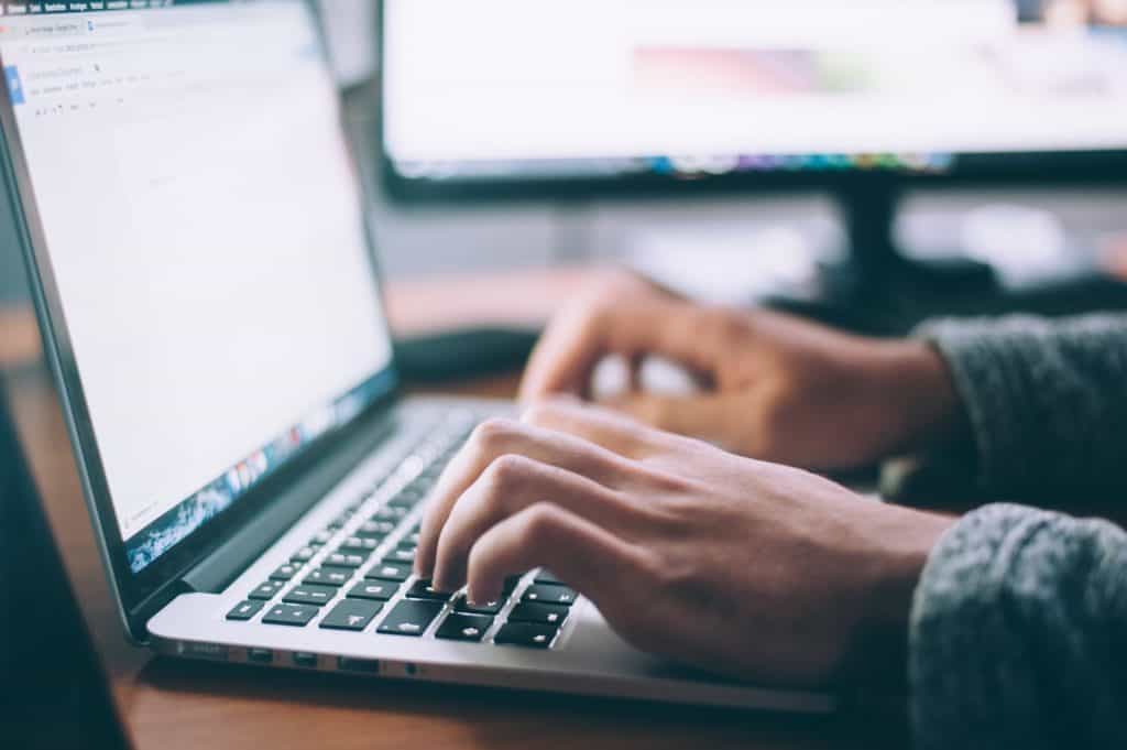 Close-up of hands typing on a laptop at a desk with a second monitor behind