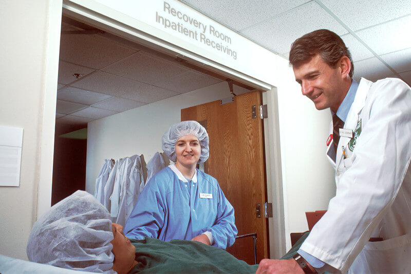 A nurse and a doctor talking beside a patient's bed outside a hospital recovery room