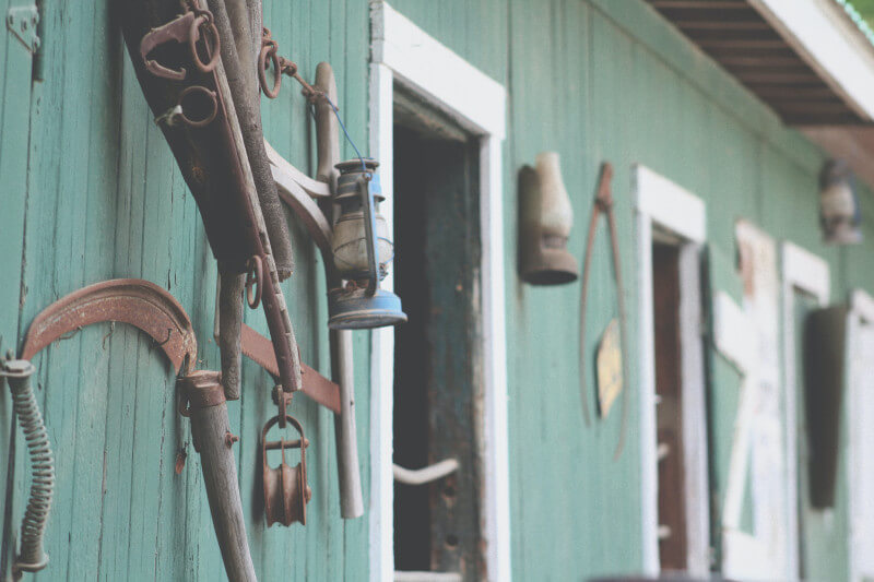 The weathered green wooden exterior of a stable, with old lanterns and tack hanging by the open doors
